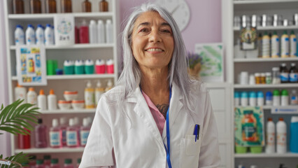 Elderly woman with grey hair smiling in a pharmacy wearing a white lab coat surrounded by various pharmaceutical products on shelves indoors