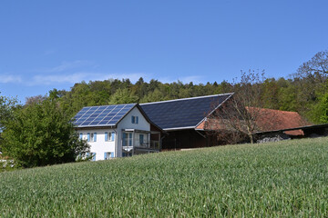 Photovoltaic or solar panels installed on the roof of farm building and family house observed in a Swiss village. There are trees and bushes around the house.