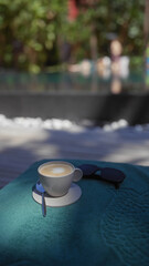 A cappuccino and sunglasses rest on a vibrant towel, suggesting leisure by a blurry poolside setting.