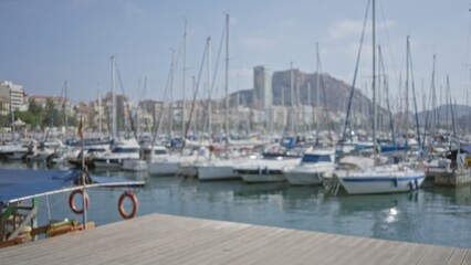 Blurred background of an outdoor marina with docked sailboats and a cityscape under a clear blue sky, featuring a defocused view of water and distant mountains.