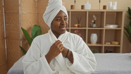 Young african woman in a spa setting wearing a white robe and towel headwrap, looking content and relaxed within a wellness center interior