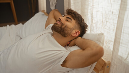 Young man relaxing on a bed in a cozy bedroom with sunlight streaming through the curtains, showcasing a peaceful indoor setting and a handsome bearded guy enjoying his personal space at home.