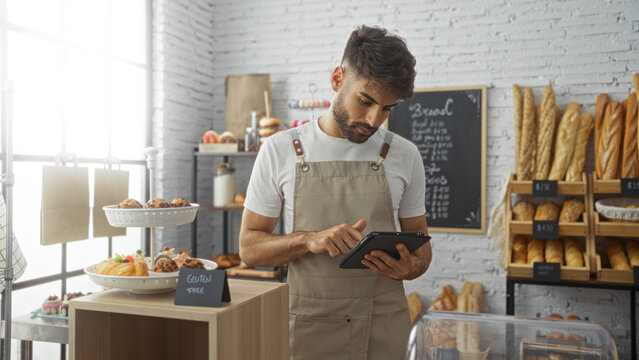 Young hispanic man working in a bakery shop using a tablet surrounded by bread and pastries - Powered by Adobe