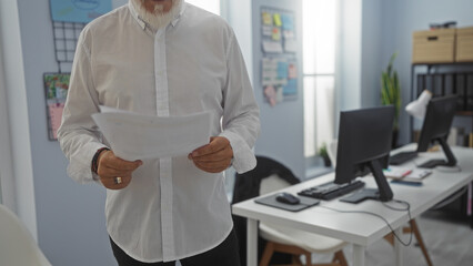 Mature man holding papers in an office setting with computers in the background and daylight...