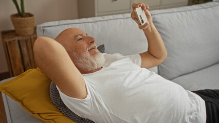 Man relaxing on sofa at home with smartphone, indoors showing grey-haired mature senior enjoying...