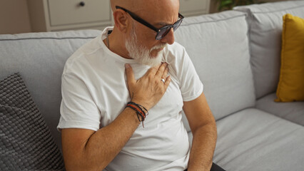 Man with grey hair sitting on sofa in living room with hand on chest, indicating discomfort or pain, wearing white shirt and glasses, showing signs of mature or senior age indoors.