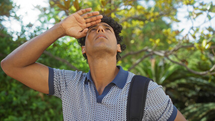 Young, hispanic, man looking up in an outdoor, urban park filled with greenery, showcasing an attractive adult wearing a polo shirt and a backpack