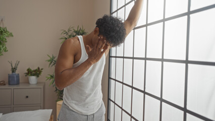 Young man feeling dizzy while leaning on a large window in a modern bedroom with plants and neutral decor