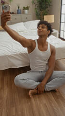 Young, hispanic, man taking a selfie while sitting on the floor in a cozy bedroom decorated with plants and neutral-colored furniture