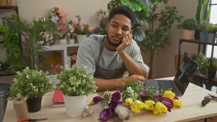 Thoughtful african man at flower shop with plants, laptop, arranging flowers, posing for portrait indoors.
