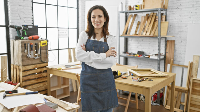 A confident young woman with crossed arms wears an apron in a well-equipped carpentry workshop indoors. - Powered by Adobe