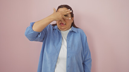 Mature hispanic woman looks uncomfortable against a pink wall, covering her face with her hand in distress.