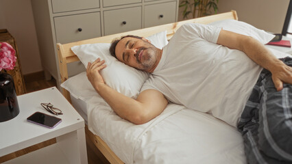 Hispanic man sleeping peacefully in a cozy bedroom at home, resting on a bed with white sheets, and a nightstand holding a smartphone and glasses.