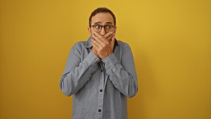 Fototapeta premium Middle-aged hispanic man covering mouth with both hands, standing isolated against a vibrant yellow background wearing glasses and a blue shirt, exhibiting a surprised expression