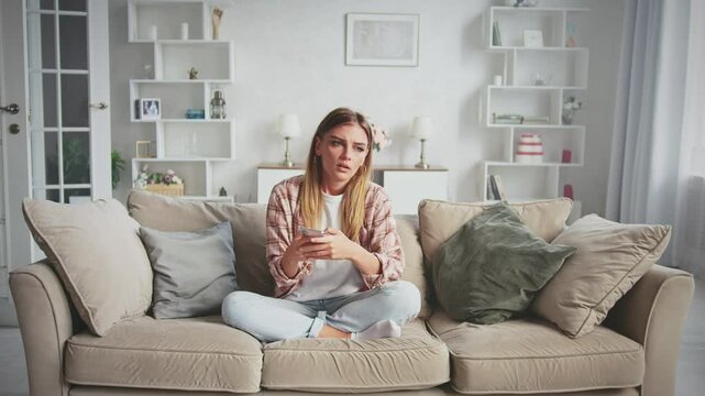 Young woman sits on a sofa in her living room, looking down at her smartphone with a sad expression. Feelings of loneliness and disappointment wash over her as she scrolls