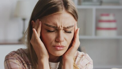 Young woman suffers from a strong headache or migraine at home. Massaging her temples and closing her eyes, she expresses pain and discomfort while feeling overwhelmed - Powered by Adobe