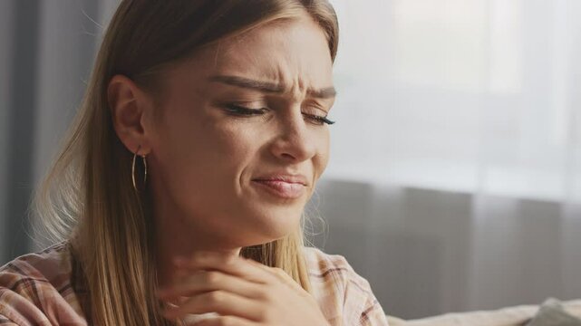 Woman touching her neck and coughing, showing discomfort from cold or flu symptoms. Signs of illness include fatigue, sore throat, and general unwellness in a home setting