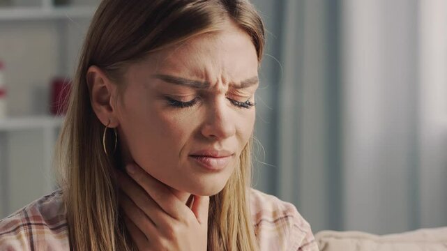 Young woman sitting on a sofa shows signs of discomfort from a sore throat. Touching her neck, she expresses pain and distress, highlighting her struggle with illness at home