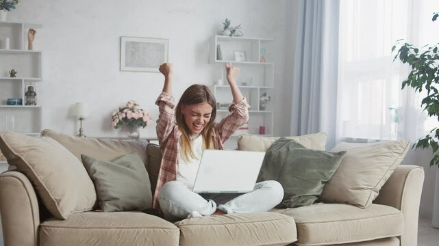 Young woman sitting on a sofa in her living room is overcome with joy as she achieves a significant victory on her laptop