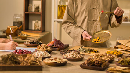In a photo depicting traditional Chinese medicine, two herbalists are shown taking notes and weighing herbal compounds to pour into wrapping paper on a table surrounded by a variety of herbs.