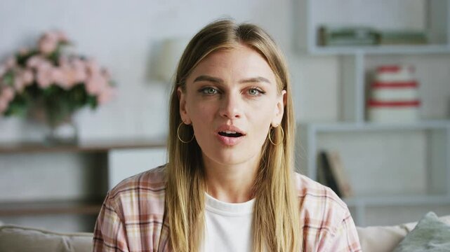 A woman with blonde hair and a pink shirt is sitting on a couch. She says hello and tells something to her interlocutor via video call.