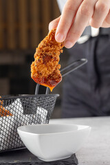 A hand is dipping a crispy, golden-brown chicken tender into a small white sauce cup. The scene is...