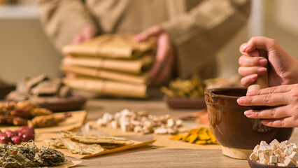 A herbalist is pounding angelica sinensis in the Oriental medication setting with a table full of herbs as another herbalist in beige clothing organizes the packets of wrapped herbal medication.
