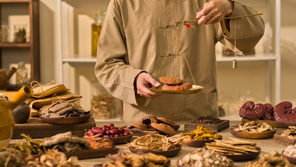 A medical practitioner weighs reishi mushrooms on a traditional scale, while other herbs and laboratory tools are arranged around the table, template picture for traditional medicine advertisements.