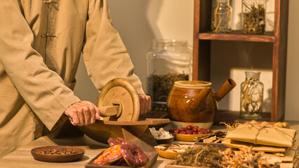 In a traditional eastern medicine room, herbs , packages, and other tools used in the field are arranged next to a herbalist who is grinding herbs with a wooden mill on a table.