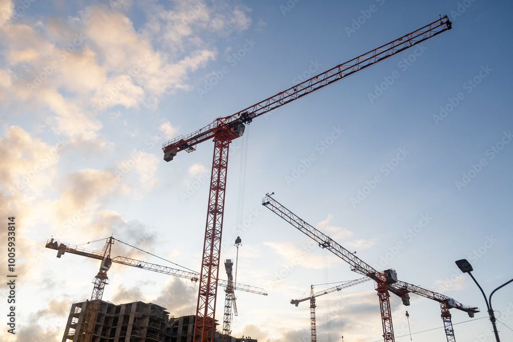 Poster cranes lift building materials at a busy construction site with an expansive sky filled with clouds 