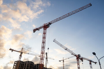 Cranes lift building materials at a busy construction site with an expansive sky filled with clouds during the afternoon