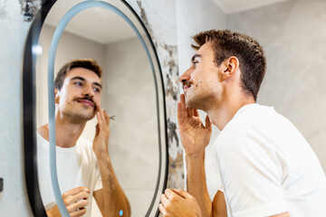 Young man trims his mustache with scissors in the bathroom in front of the mirror.