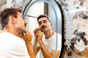 Young man trims his mustache with scissors in the bathroom in front of the mirror.
