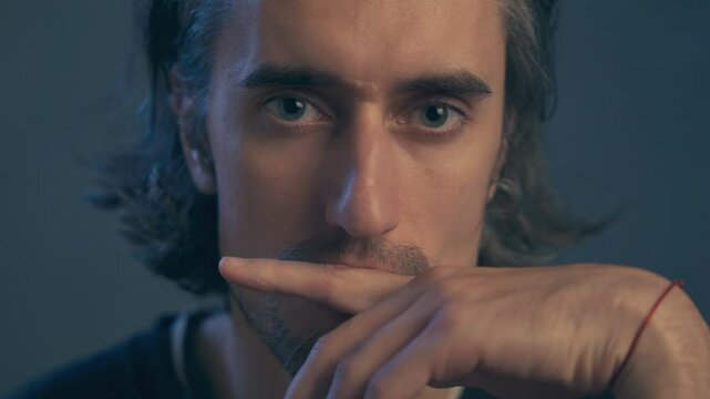 a young man with long hair, large expressive eyes, European appearance, looks intently, raises his eyebrows questioningly, looks incredulously into the frame, covers his mouth with his hand