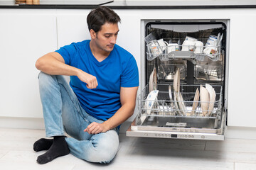 Tired and bored man putting dirty plates in dishwasher machine in the kitchen. Household and exhausting cleaning day concept