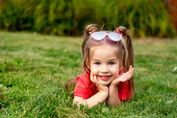 a child girl on a green lawn, a happy little child in glasses and a red T-shirt in summer on the grass rejoices and laughs, a place for text