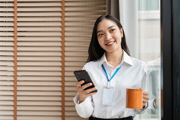 Young Professional Woman Holding Smartphone and Coffee Mug in Modern Office Setting