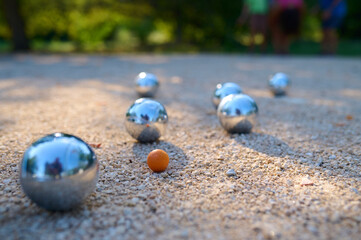 Close-up of petanque balls scattered on gravel with the target ball in focus. The shiny metal balls reflect sunlight, capturing a playful outdoor moment in a park setting