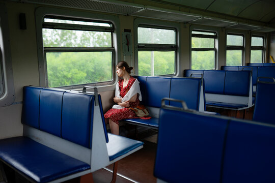 Mid adult woman in a red dress riding a train, looking out the window during daytime.