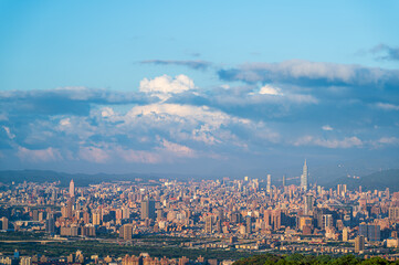A breathtaking panoramic view of a bustling city at sunset. Soft, fluffy clouds drift across the sky, casting a warm glow over the urban landscape. Captured in Dading Mountain, Shulin, Taiwan.