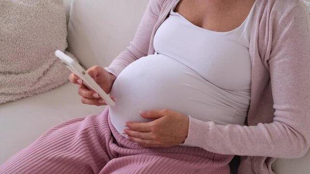 Close up shot of a pregnant middle aged Caucasian woman using mobile phone for online entertainment in cyberspace and caressing tummy at home.