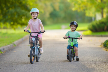 Happy smiling sister and little brother with helmets running and riding on bikes on road at town...