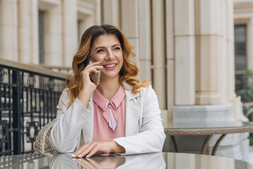 Woman Phone Call Cafe - Smiling woman talking on her phone while sitting at a table outside a cafe.