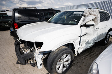 A white SUV with extensive damage to the front and side sits in a parking lot, likely after a collision. The airbag is deployed, indicating the severity of the accident.