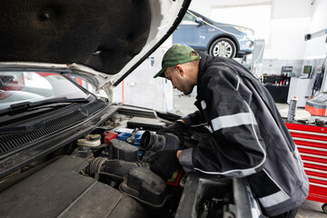 A focused mechanic inspects a car engine in a modern auto repair shop. The garage has tools and...