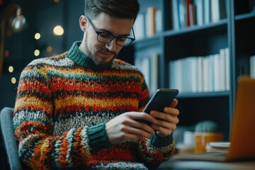 Fototapeta premium Man wearing colorful sweater working on smartphone in a professional environment, surrounded by books and a laptop.
