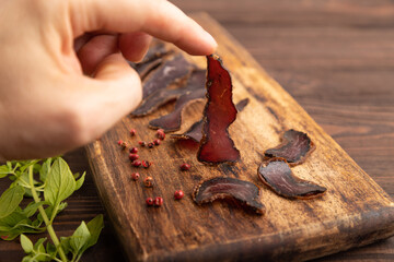 Armenian Basturma dried meat with hand on wooden cutting board with pepper and herbs on brown wooden. Side view, selective focus.