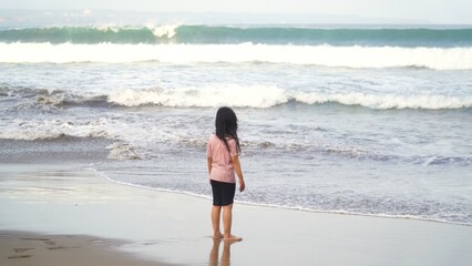 An Asian girl with long black hair is standing facing the sea wearing a pink t-shirt and black shorts on a cloudy morning.