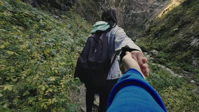 First person view of a young woman with a backpack leads by the hand during a walk together on the mountain trail between dense grass. Concept of active outdoor hike tourism