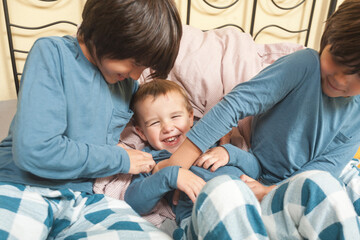 Three boys in the same blue pajamas play together in bed and tickle their little brother.